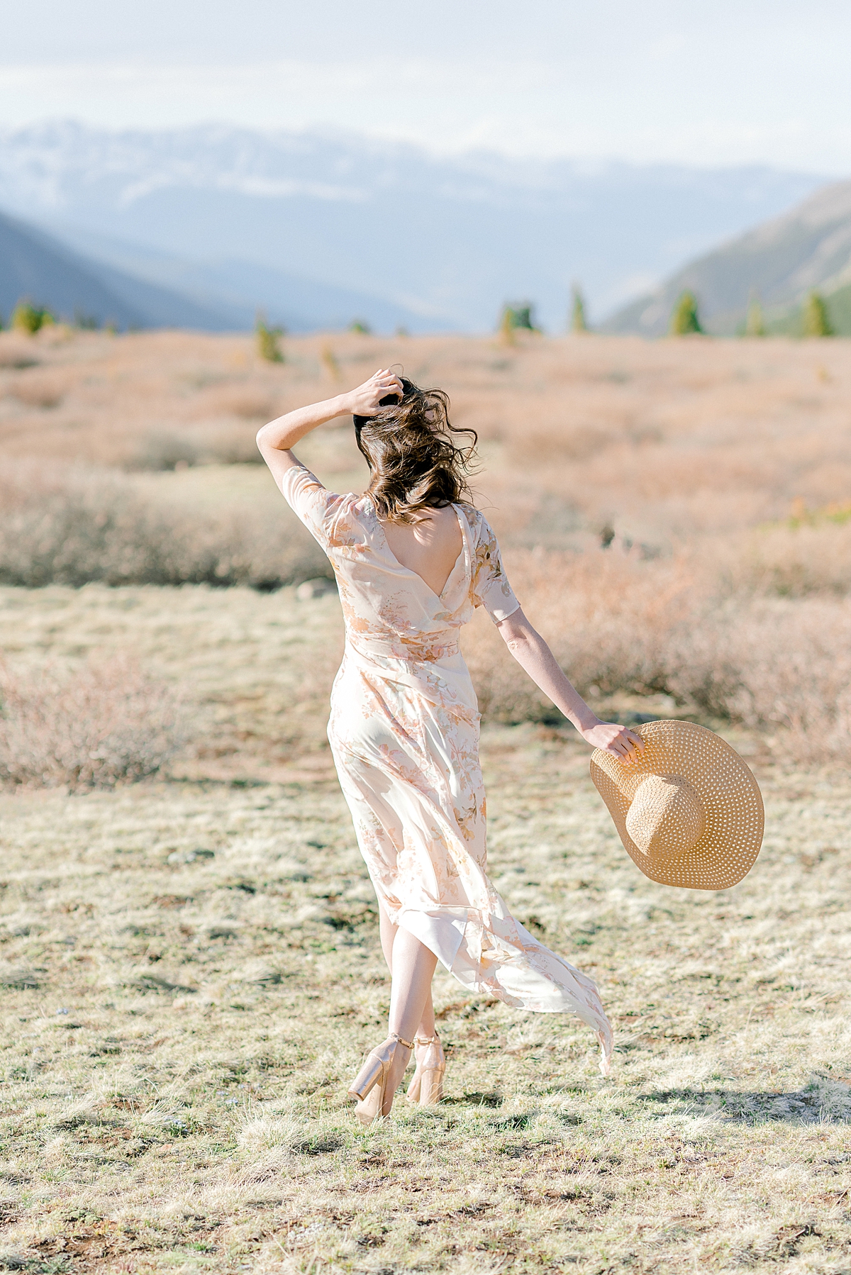 a girl in a peach floral dress and sunhat walks towards the mountains