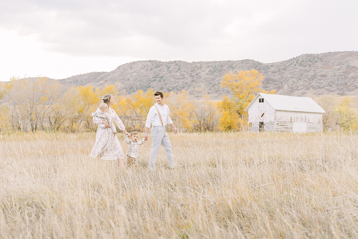 A sweet family of 4 walks through a field with a whitewashed barn in Littleton, Colorado for Denver Mountain Family Photos.