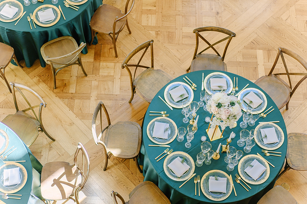 An aerial photograph of wedding reception with green linens, x-back chairs, gold gilded glassware, gold flatware, and candles.