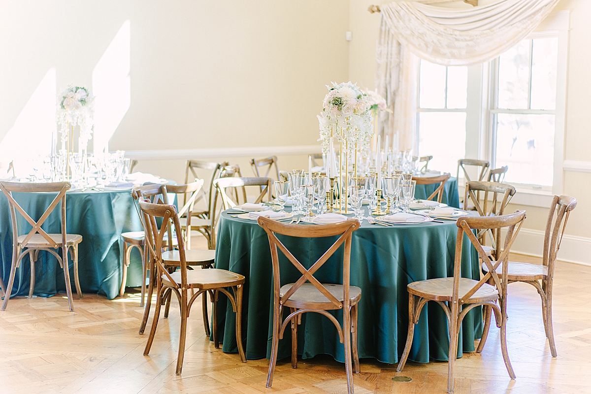 Photo of a wedding reception tablescape featuring green linens, x-back chairs, gold gilded glassware, gold flatware, a spruce twig tucked into every napkin, and a palette of neutral florals.