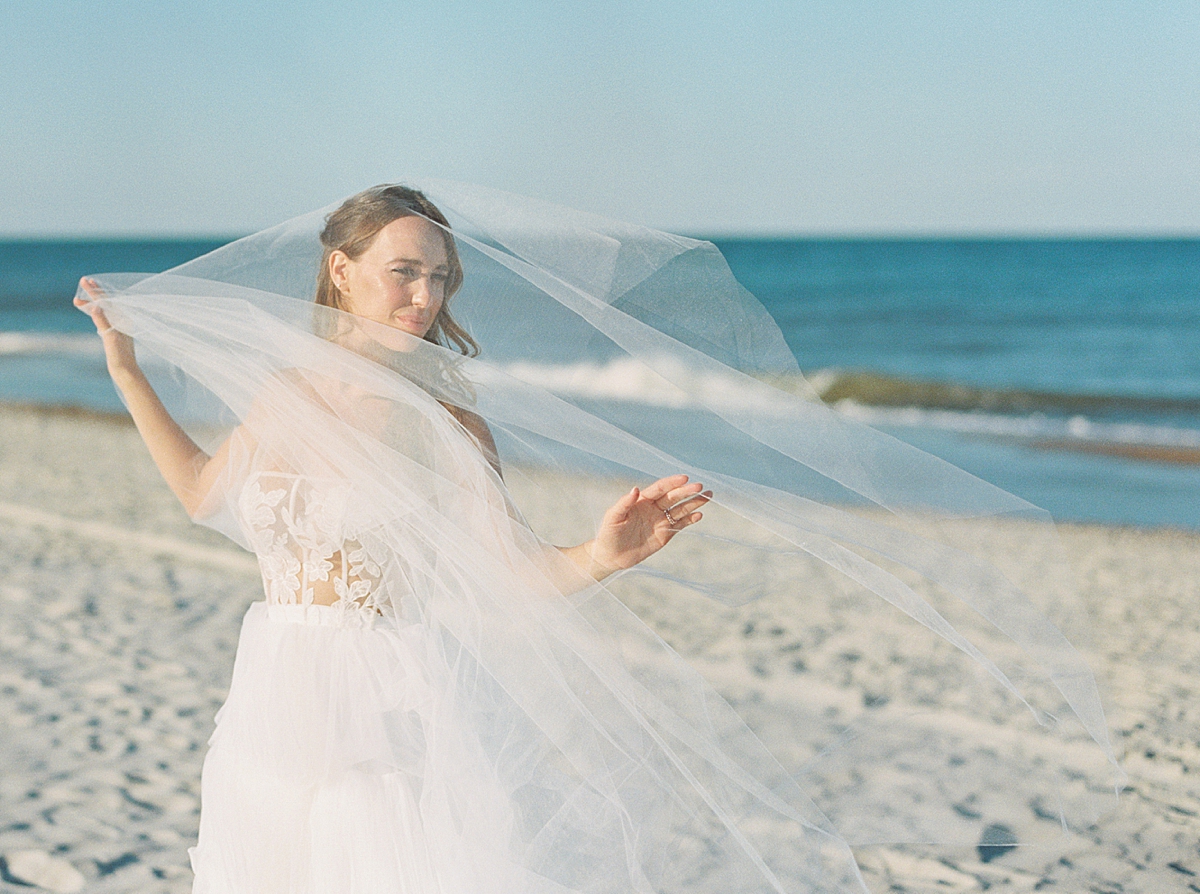 Bride at the beach while her veil blows in the wind. Veil is from Madeleine Fig. Wedding Dress is by Elizabeth Leese.