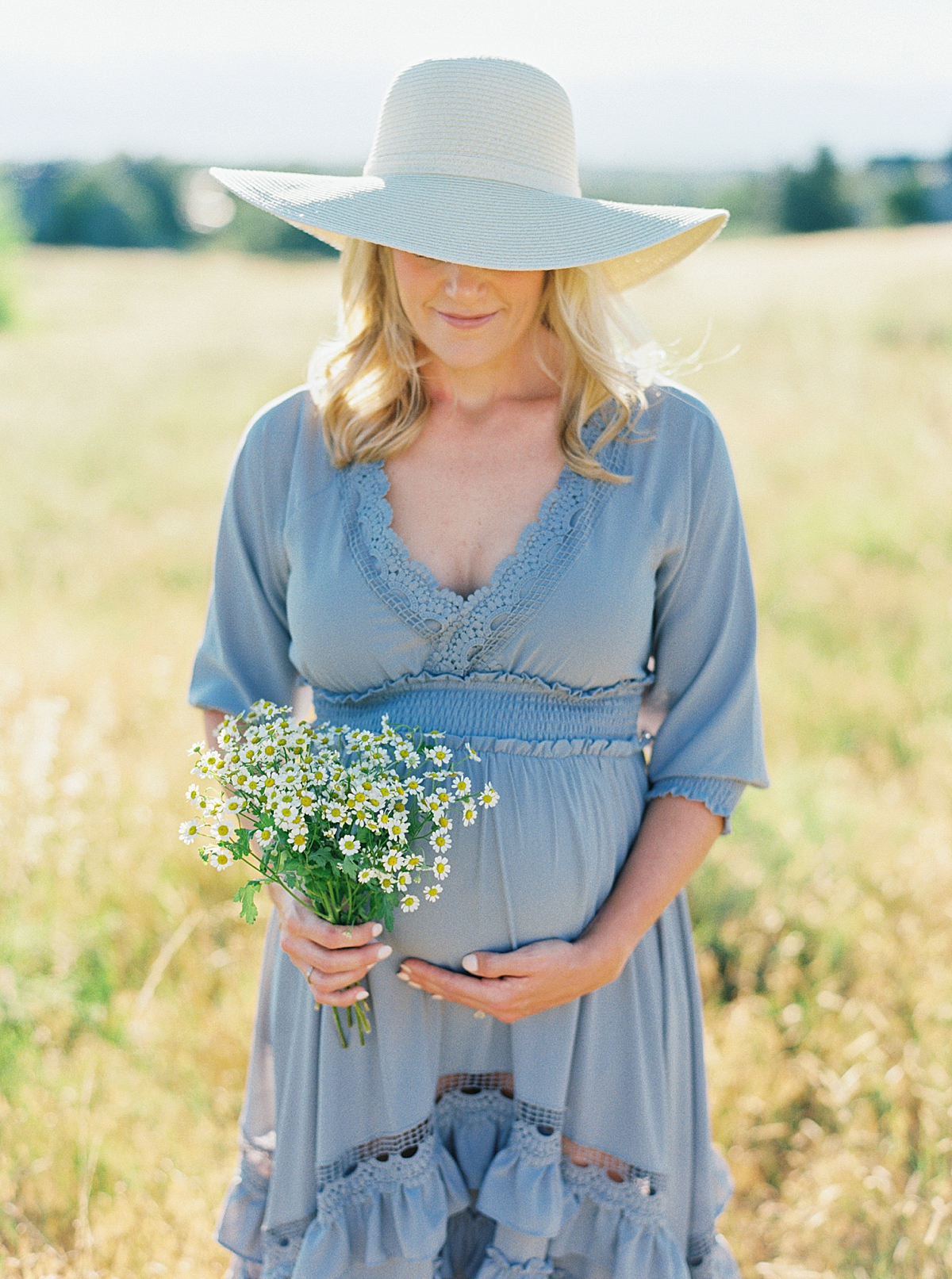 An expecting mom wears a straw hat and holds a bouquet of wildflowers in a field.