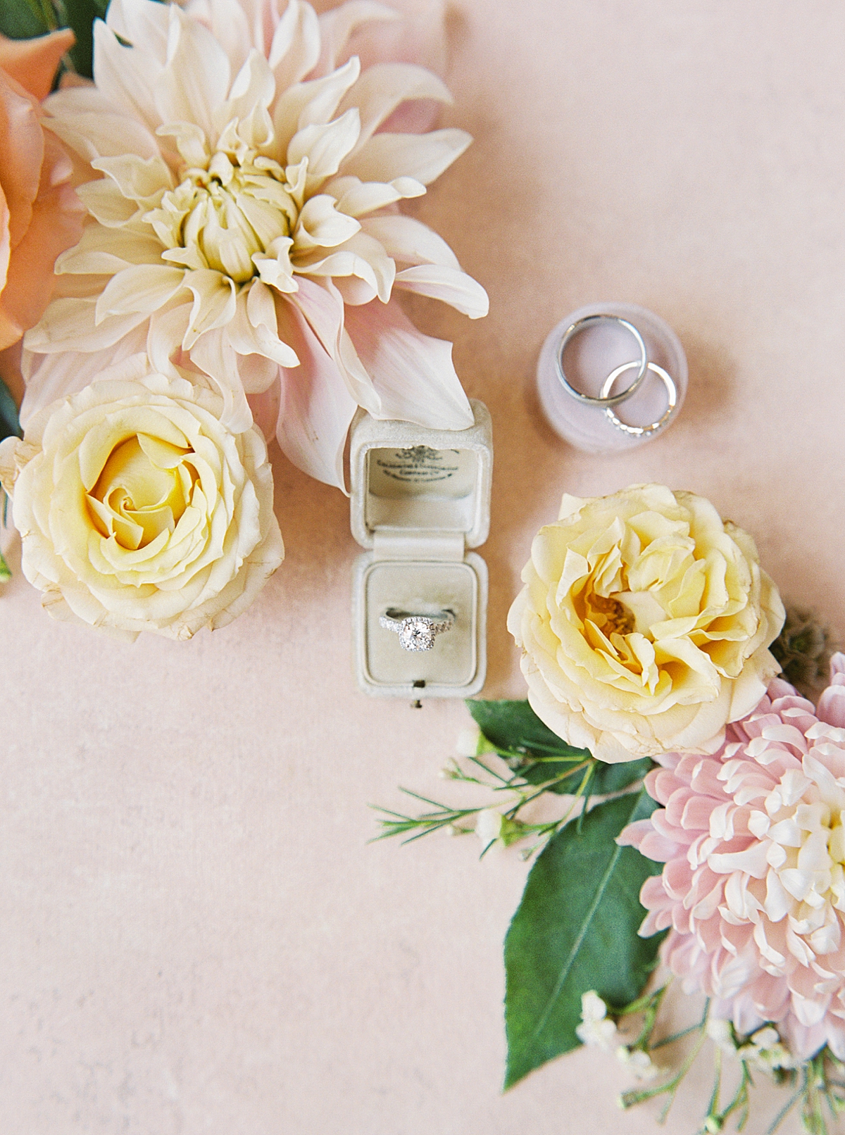 A flat lay on a pink Chasing Stone surface, with an engagement ring in a cream velvet vintage ring box. Surround by cafe au lait dahlias and roses.