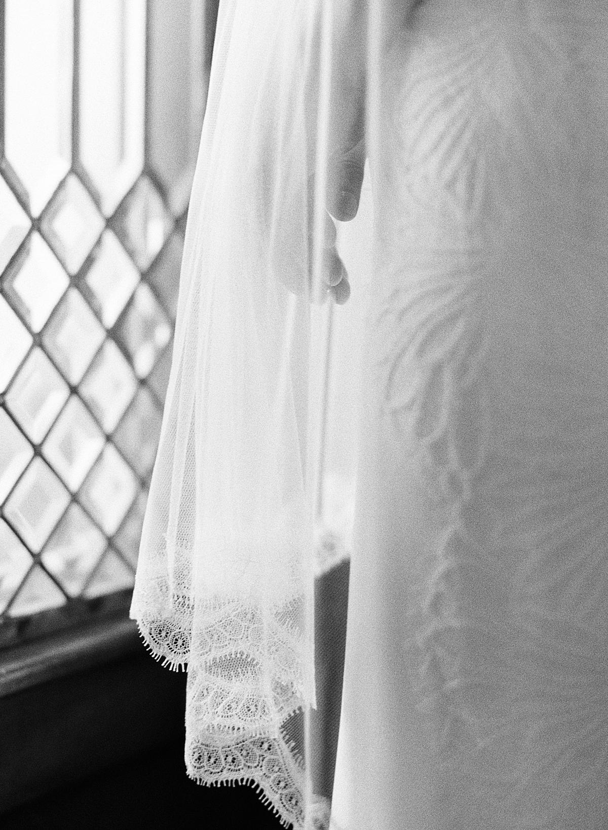 A bride gazes out the window with a lace veil covering her fingers, photographed on Ilford Delta 3200 black and white film.