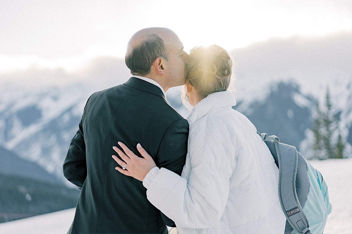 A groom kisses his bride on the head while they look at the snowy mountains in Aspen, at The Little Nell overlook.